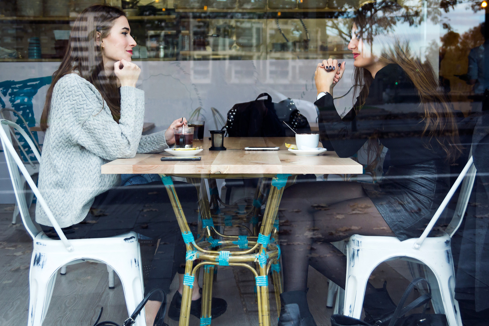 Two women seated across a café table, seen through a window with reflections.
